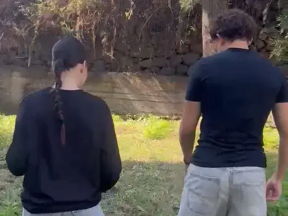Due ragazzi ripresi di spalle camminano su un prato all'aperto, davanti a un muro di pietra a secco e alla vegetazione